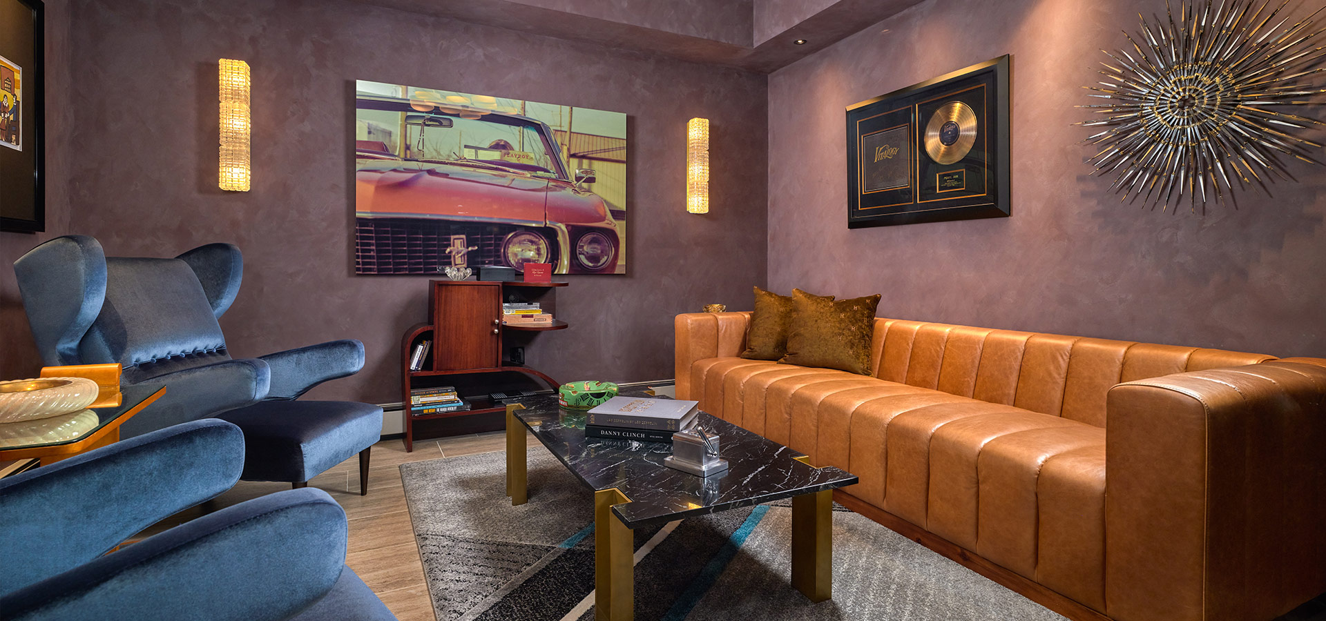 Pirela Atelier. Glamorous mid-century modern lounge with a tan leather sofa, blue velvet chairs, and a sputnik chandelier against mauve textured walls.