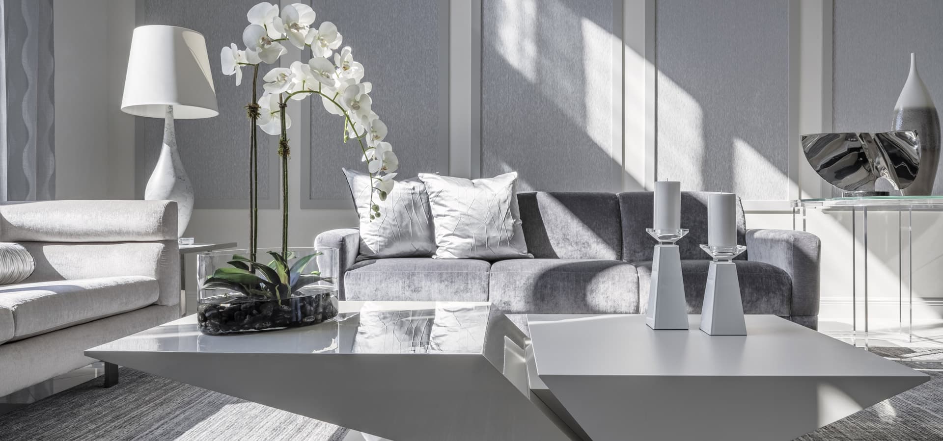 Pirela Atelier. Luxurious monochromatic living room featuring a silver velvet sofa, modern geometric white coffee table, and textured gray wall panels.