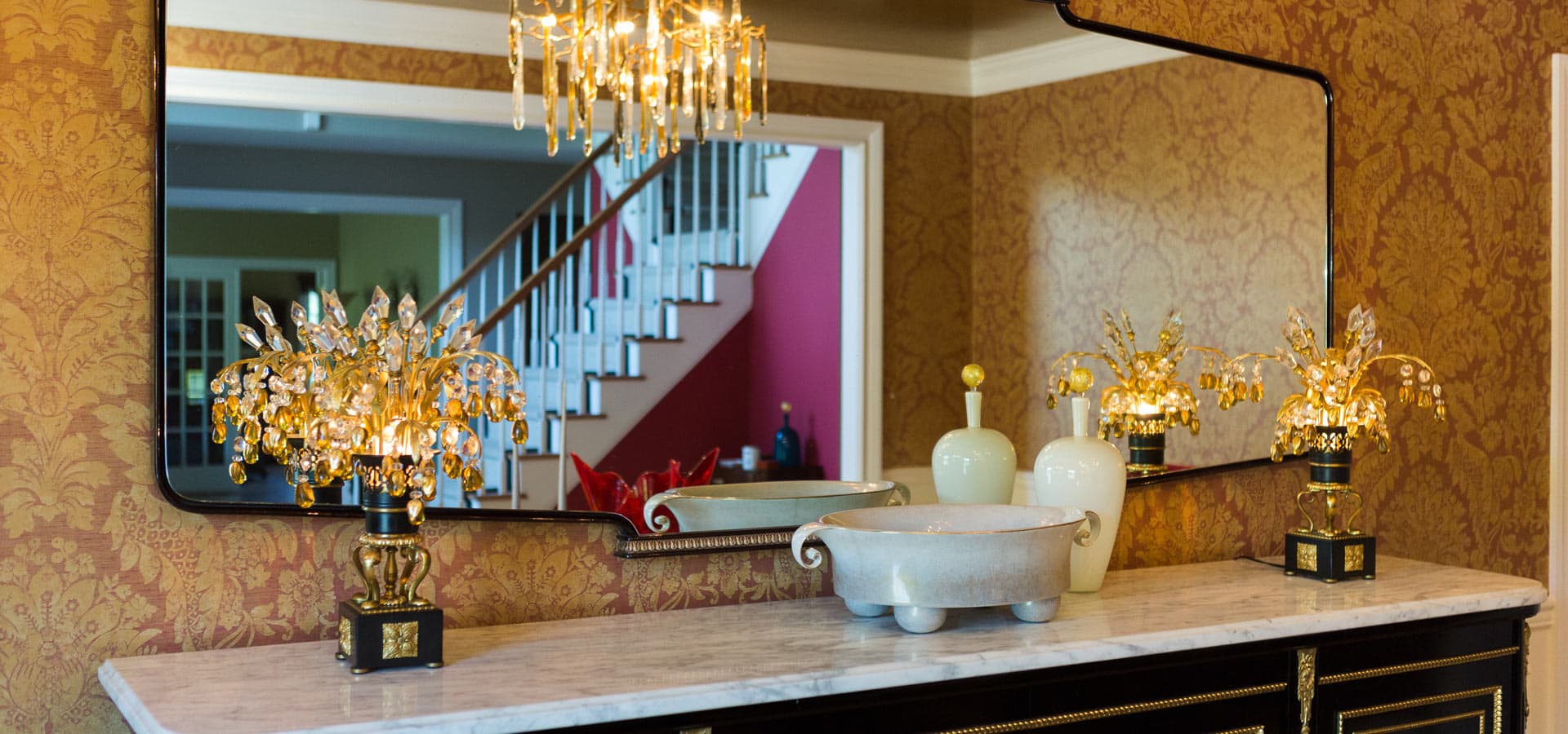 Pirela Atelier. Traditional luxury decor detail with crystal and gilt lamps reflecting in a wide, rectangular mirror.